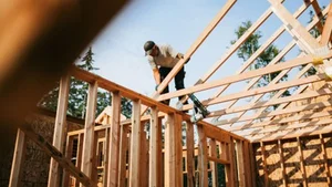 A person stands on wooden framing at a construction site, installing roof beams on a partially built structure with exposed studs and trusses; sunlight illuminates the light-colored lumber, and trees and sky are visible beyond the unfinished walls.
