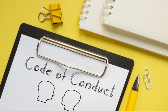 Clipboard holding a sheet of paper with text that reads Code of Conduct above two simple outline profiles of human heads facing each other. The clipboard sits on a bright yellow surface alongside office supplies including a yellow pen, a paper clip, a binder clip, and two spiral notebooks partially visible in the upper corner.