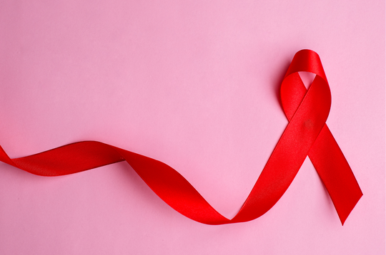 A red awareness ribbon lies on a soft pink background, with one end forming the familiar looped ribbon shape on the right and the remaining length extending loosely to the left in a gentle curve.
