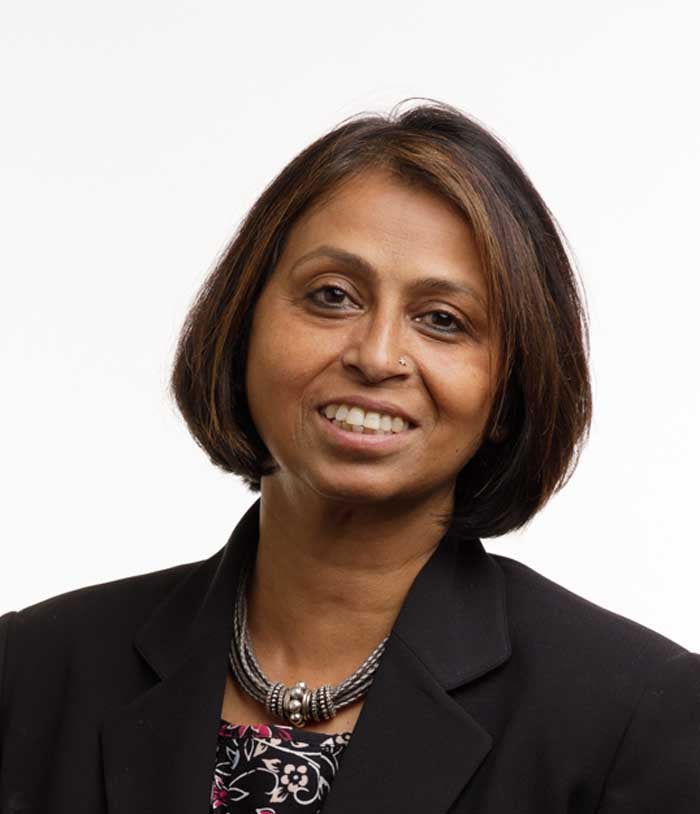 Mahasweta Girgenrath, a woman with brown hair, wears a black blazer and a black floral shirt on a white background.