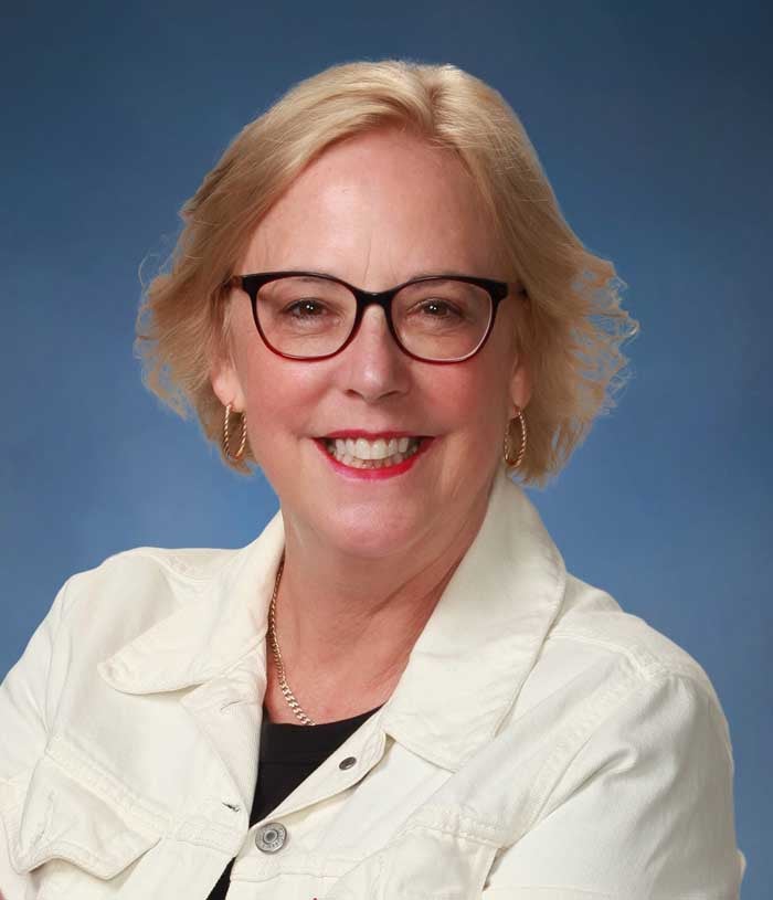 Susan Howlett, a woman with short blonde hair, wearing a white denim jacket on a blue background.
