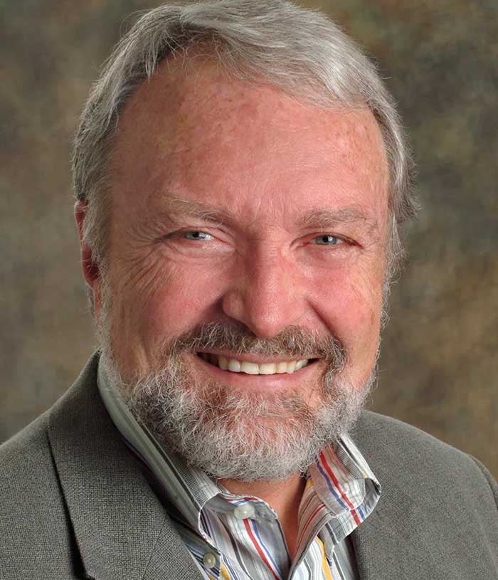 Steven Cummings, a man with gray hair and beard, wears a gray blazer and multi-colored striped shirt on a brown background.