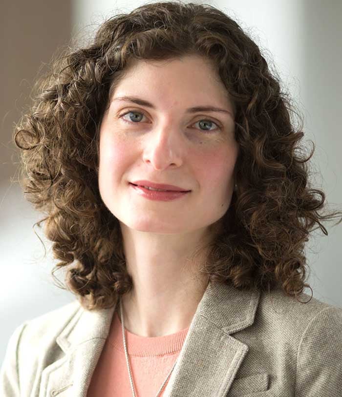 Sofiya Milman, a woman with brown curly hair, wears a tan blazer and a peach sweater.