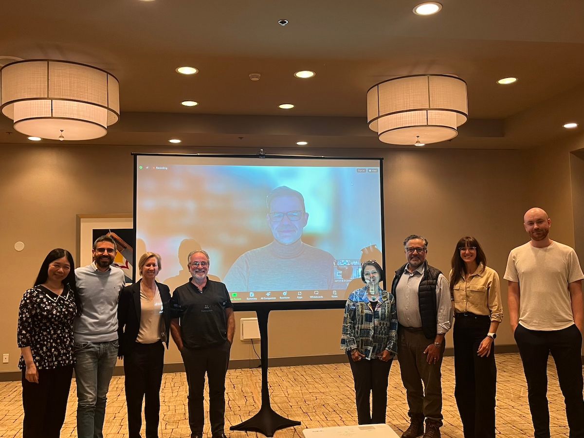 The FSHD Bonus Prize judges stand together both in-person and virtually with XPRIZE EVP, Health Jamie Justice, PhD. One judge is on Zoom on a project screen while the rest stand on either side of him in a conference room.