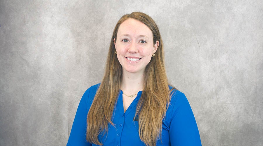 Maren Price - Woman with long straight light brown hair, wearing a bright blue blouse and small earrings, smiling in front of a light gray textured background.