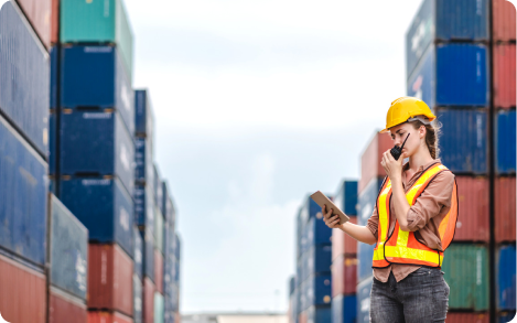 Woman in safety gear using a tablet among shipping containers.