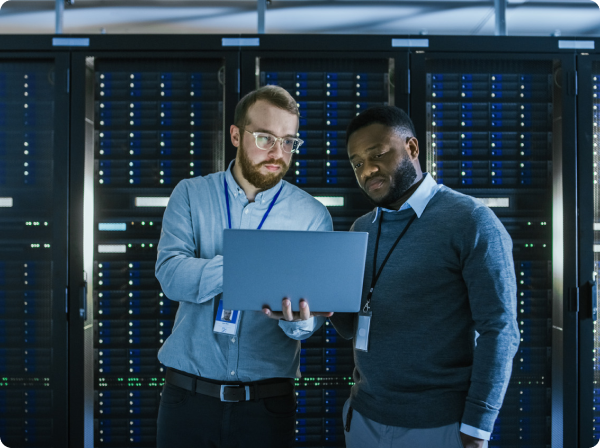 Two men reviewing data on a laptop in a server room with racks of servers.
