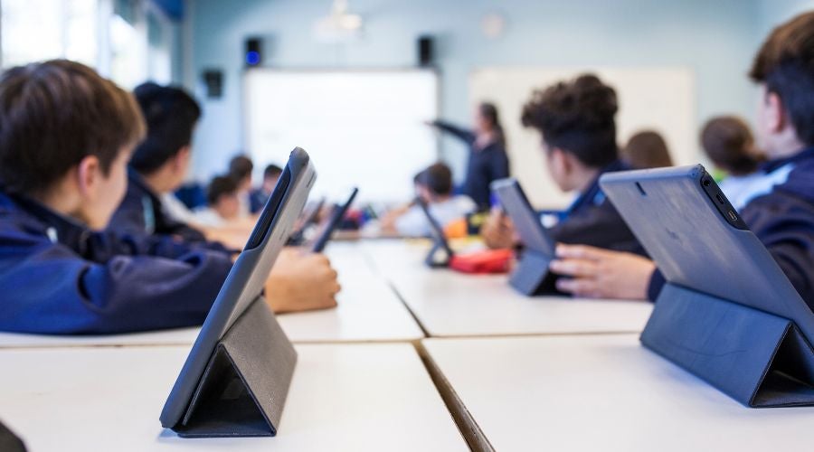 Students in a classroom using tablets on desks, with a teacher pointing at a screen.