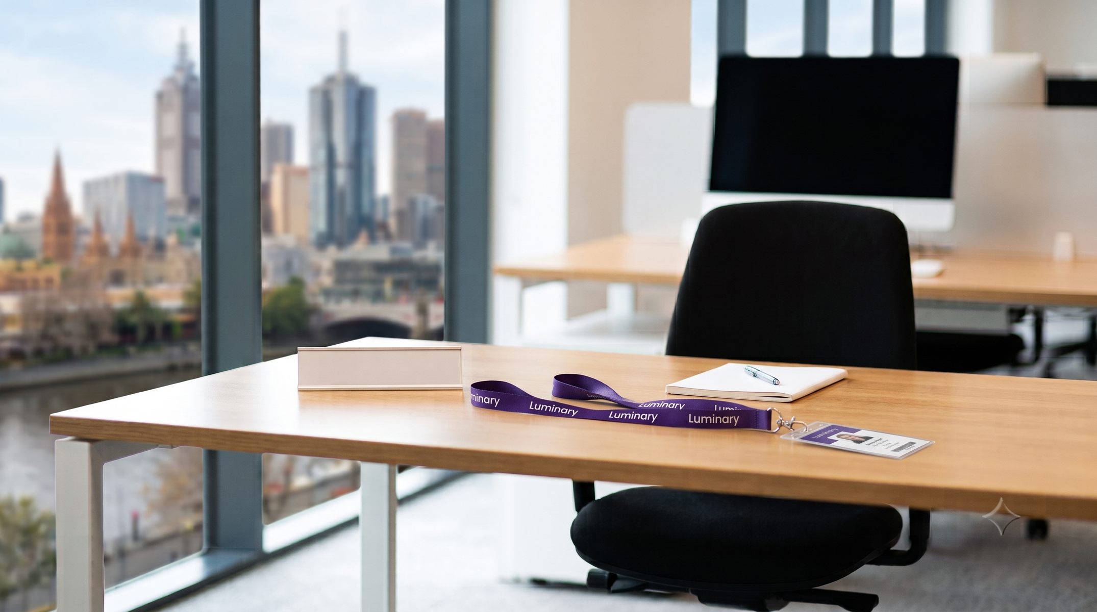 A desk set up and waiting for a new starter, with a Luminary lanyard, overlooking Melbourne. Generated by Gemini.