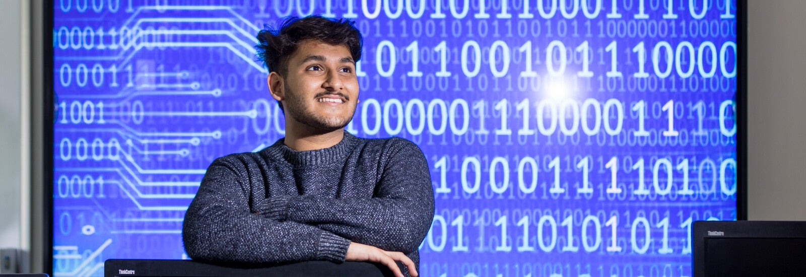 A student leaning on a computer, smiling, in front of screen