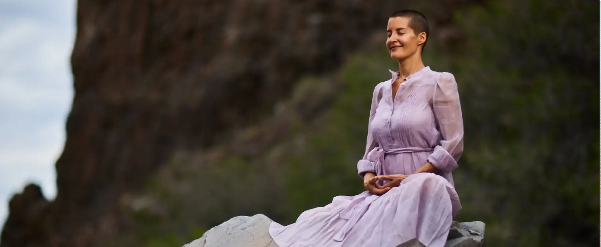 A woman stands peacefully with her eyes closed, enjoying a breeze on a mountain-top
