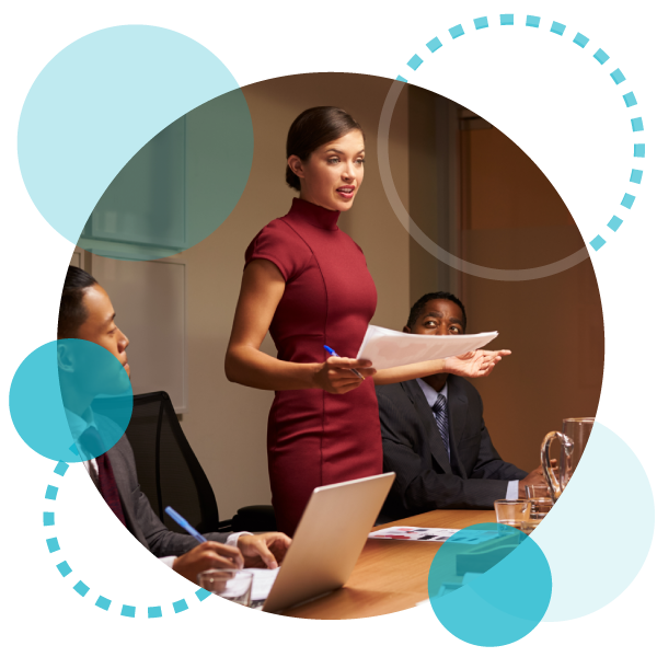 confident woman leader standing in a board room holding papers and a pen surrounded by two of her colleagues who are sitting?auto=format&q=75