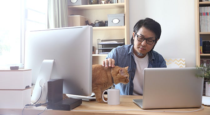 a man sat at a desk whilst stroking a cat that is on the desk