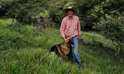 man walking through a field smiling at the camera whilst touching his dog