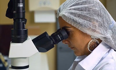 a woman in a lab with a hair net on looking into a microscope