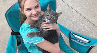 a young girl smiling whilst sat on a deck chair holding a cat with both looking at the camera