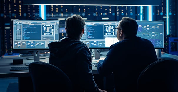Two people observing data on multiple computer screens in a dark room.