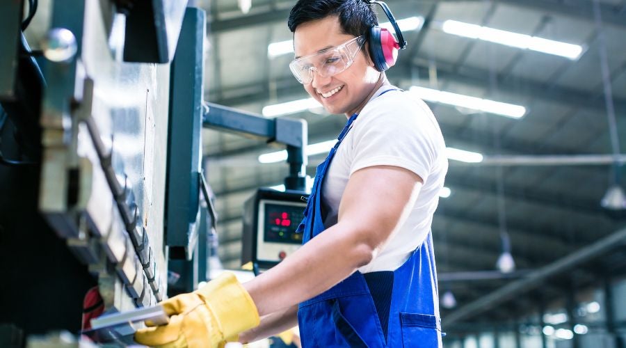 Factory worker wearing safety goggles, ear protection, and gloves operates machinery in an industrial setting, smiling while working.