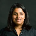 Picture of Luminary employee, Pooja smiling at the camera against a black wall