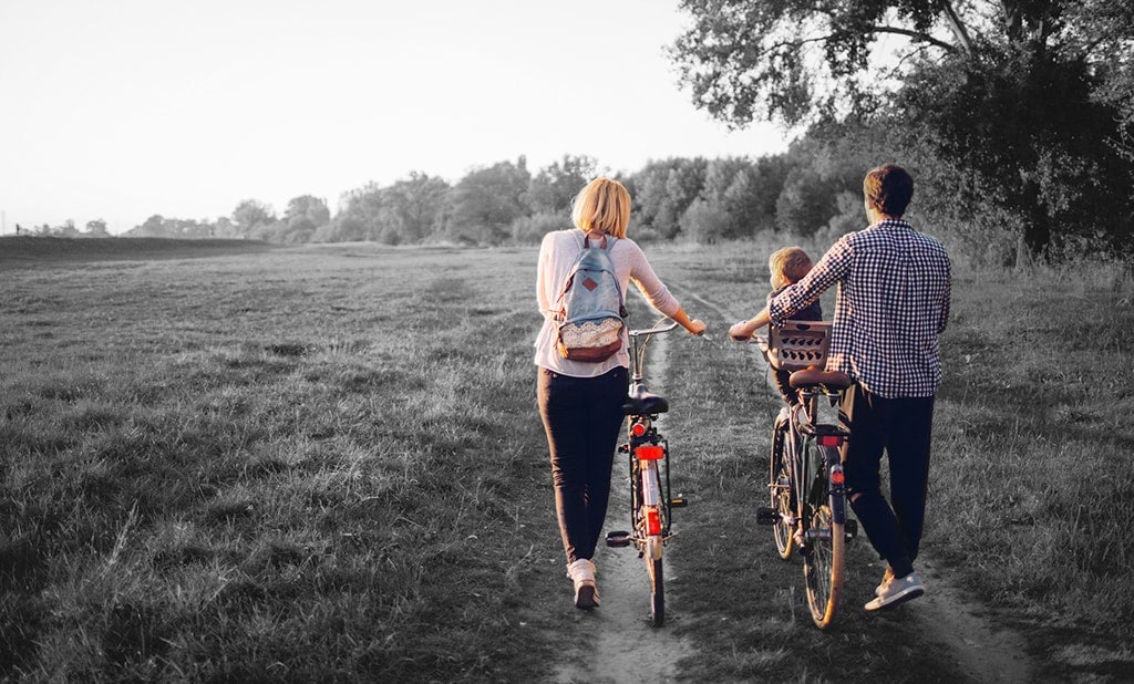 A couple walking their bikes with their child sitting in one of the bike baskets.