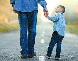 Parent walking down the street with a small child holding their hand and looking up at them.