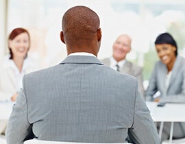 An image of a man in a suit facing away from the lens, talking to a table of people.