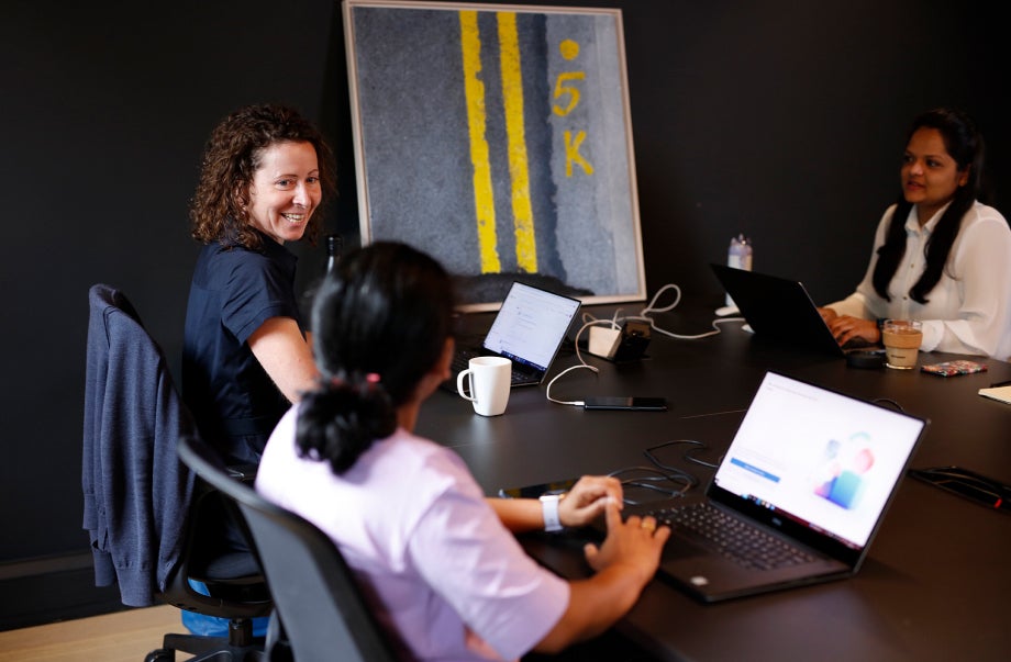 Three individuals are sitting down at a work desk with their laptops in front of them. They are smiling and talking to one another.