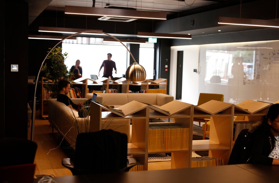 Individuals are in the All human office and are at their desk. There are open shelves and a long modern couch with an individual working on their laptop.