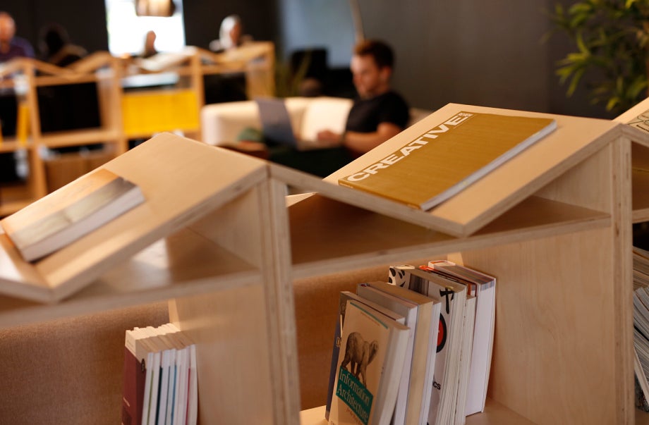 An open shelf with design books on it is in the foreground and background. In the centre of these shelves there is an individual using their laptop.