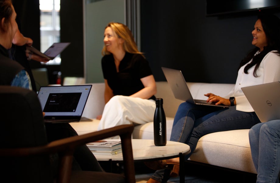 A group of people are sitting down in armchairs or on a long white couch. While their laptops rest on their legs, they are smiling and talking to one another.