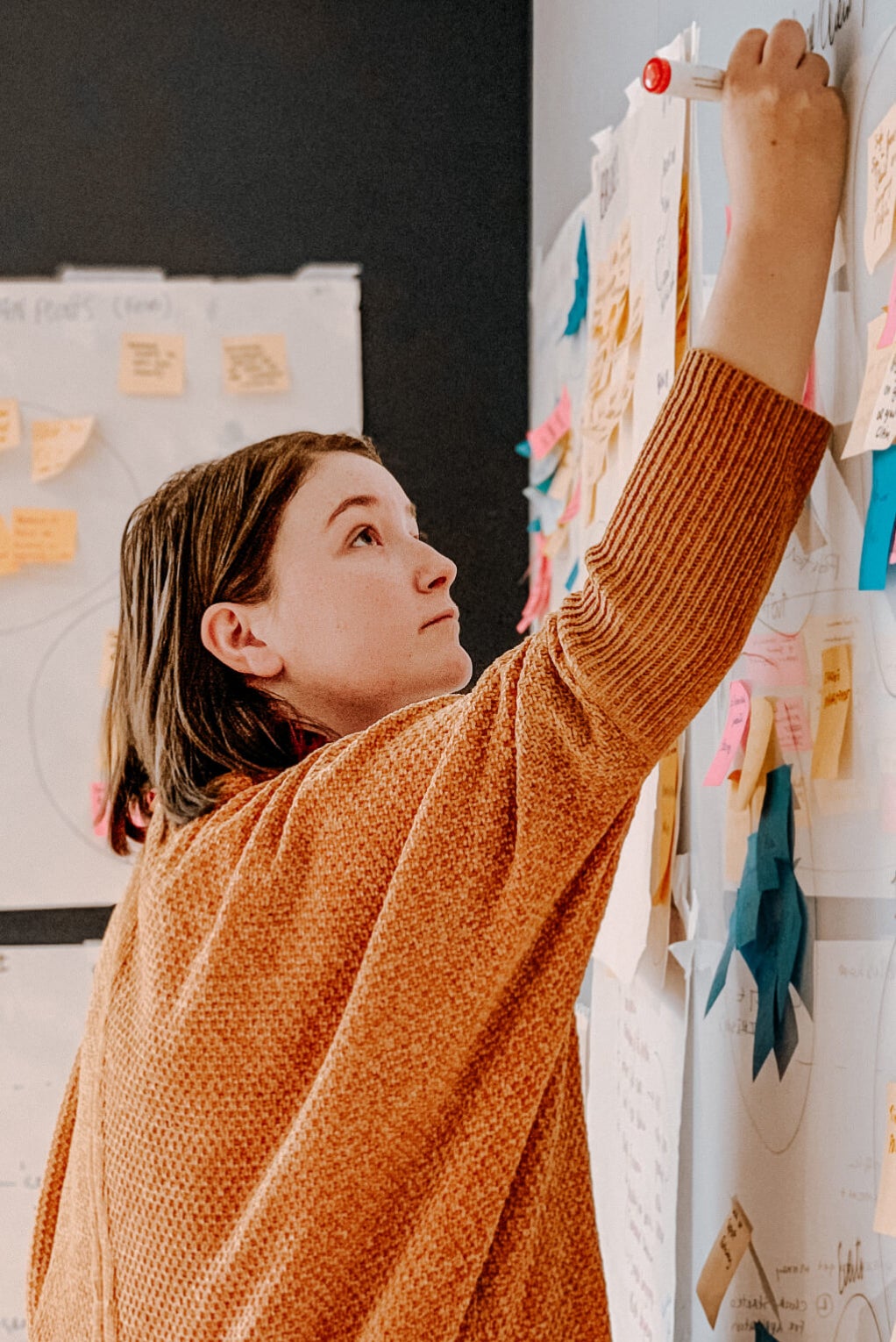 An individual is writing on a sticky note with a whiteboard maker which is placed high on the white board.