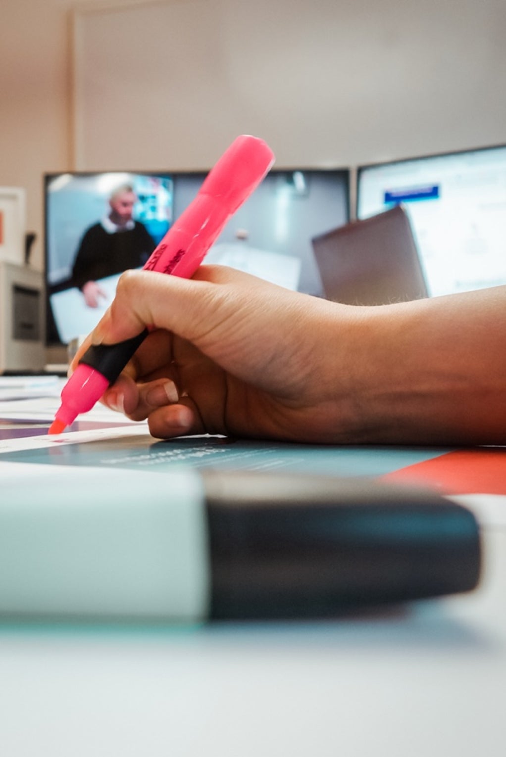 A close up of a hand holding a pink highlight marker as they are about to write something down. Another marker is placed in the foreground.